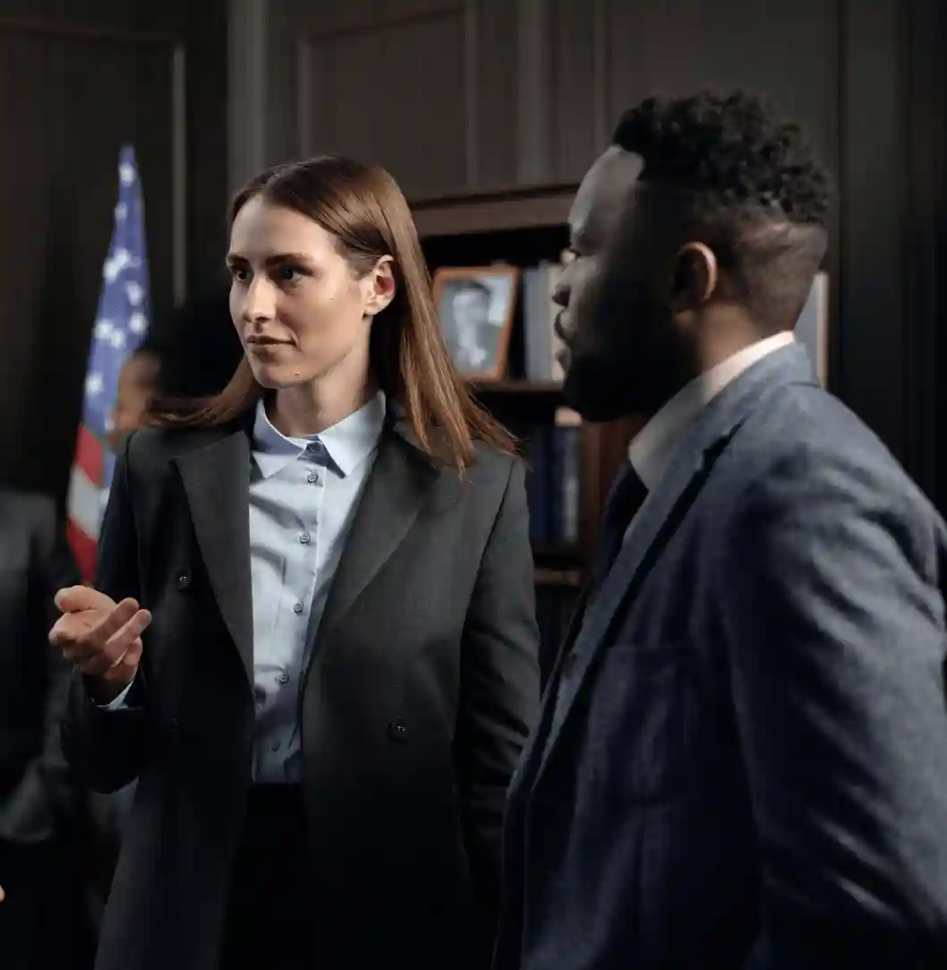 A woman in a gray suit talking to a man in a blue suit in a professional setting, with an American flag in the background.