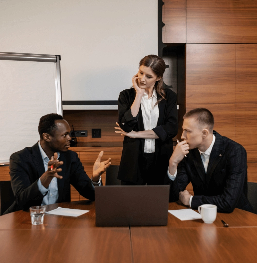 A group of three professionals having a discussion in a meeting room, with a laptop on the table and a flip chart in the background.