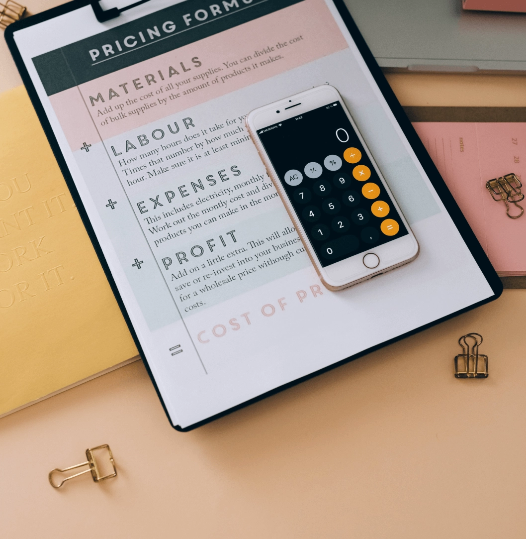 A calculator app open on a smartphone placed beside a pricing form on a clipboard, with other stationery items like a notebook, paper clips, and a binder in a light-colored setting.